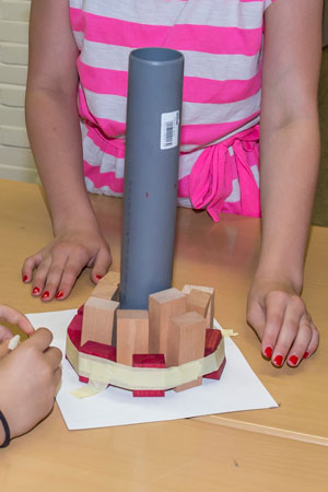 Kinderhände bauen einen Turm aus Holzklötzen und einem grauen Rohr auf einem Tisch.