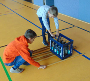 Zwei Kinder rollen eine Getränkekiste auf Holzstäben über den Boden, um Reibung zu verringern.